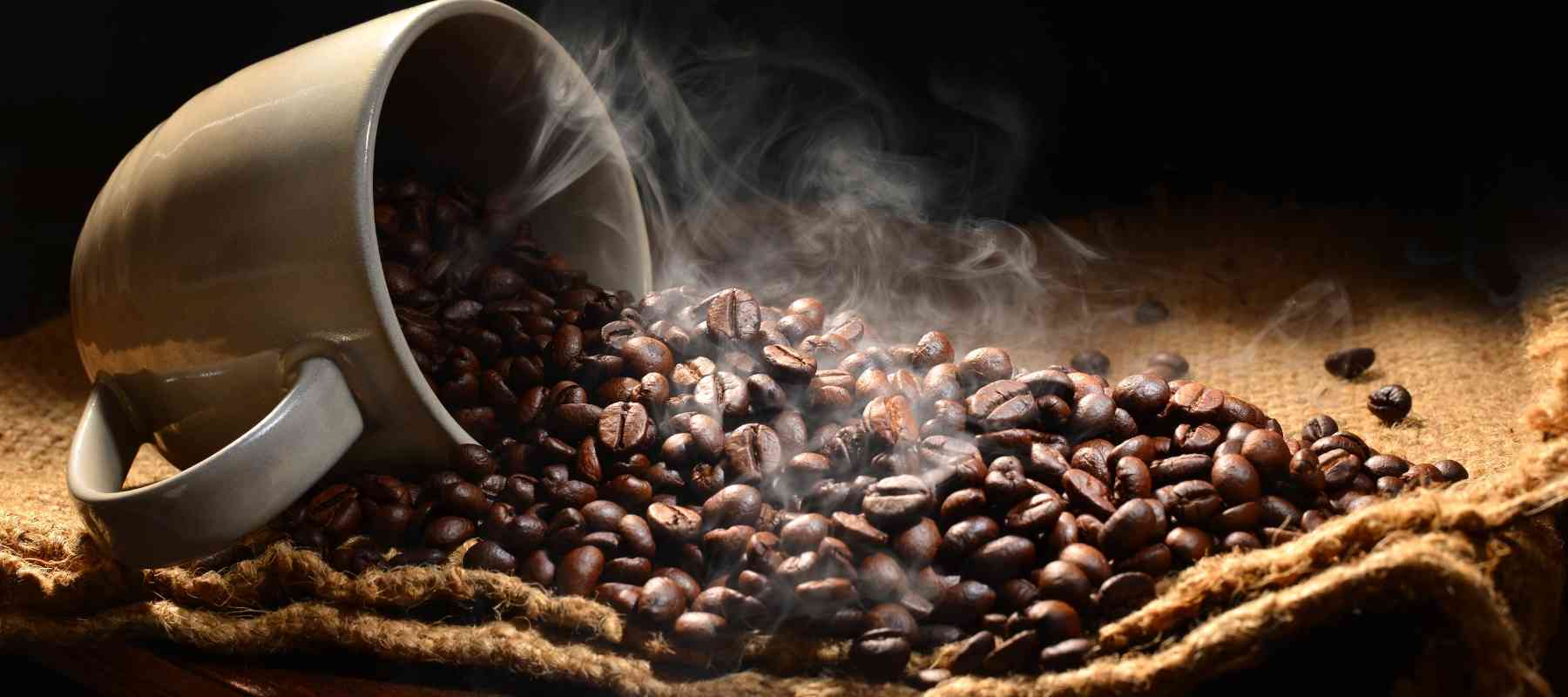 White mug spilling coffee beans onto a burlap surface with steam rising.